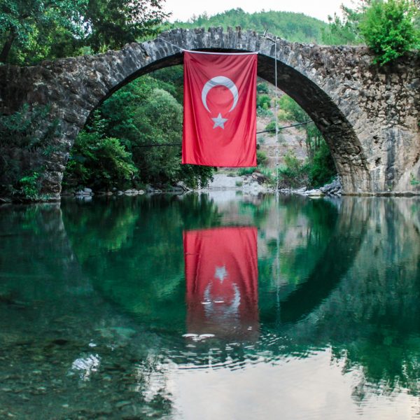A serene view of a stone bridge with a Turkish flag reflecting on the water below.