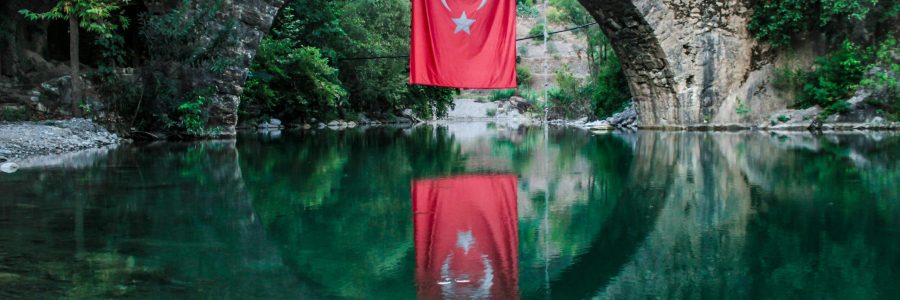 A serene view of a stone bridge with a Turkish flag reflecting on the water below.